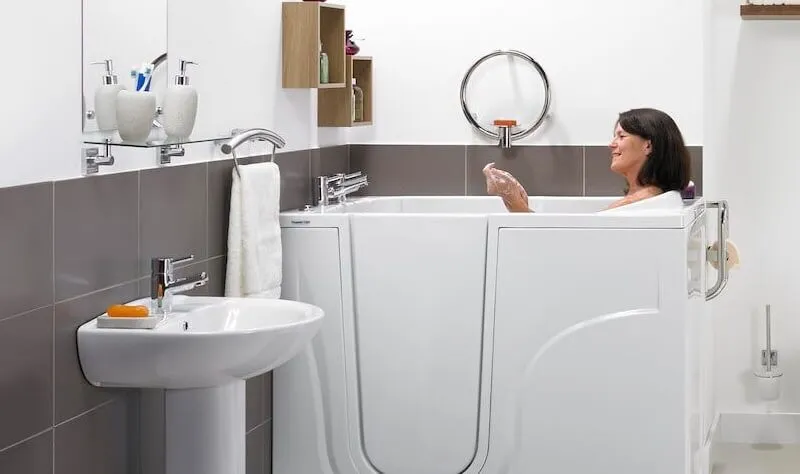 A woman relaxes in a modern walk-in bathtub in a bathroom with gray and white tiles, a wall-mounted sink, and shelves holding toiletries and towels.