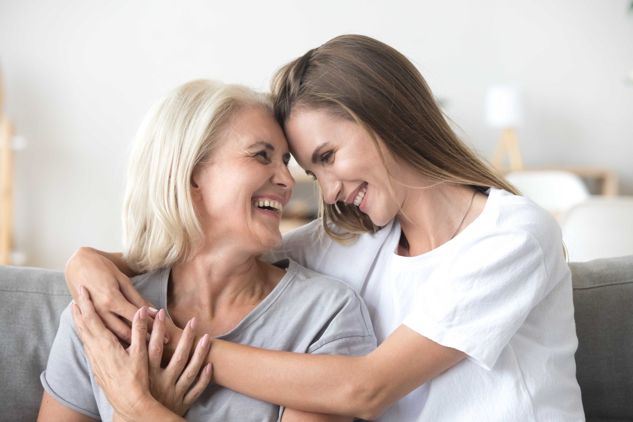 Two women, one older with gray hair and one younger with long brown hair, sit close together on a couch, smiling and embracing each other affectionately in a bright, cozy room.