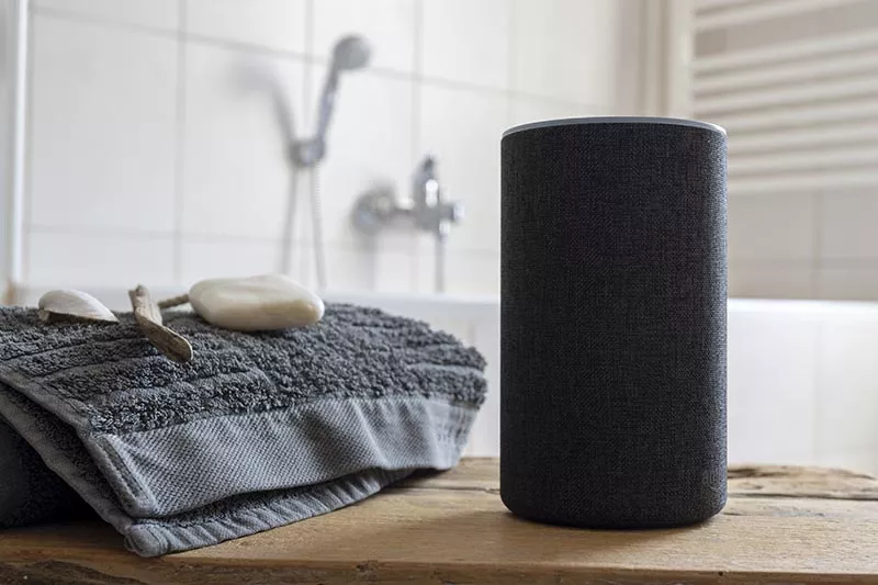 A smart speaker sits on a wooden surface next to folded gray towels, a toothbrush, and soap, with a bathtub and showerhead visible in the tiled bathroom background.