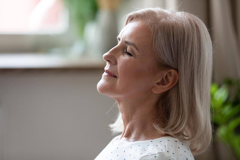 An older woman with light gray hair sits indoors with her eyes closed and a peaceful expression, appearing relaxed and content. Soft natural light and a blurred background with green plants are visible.