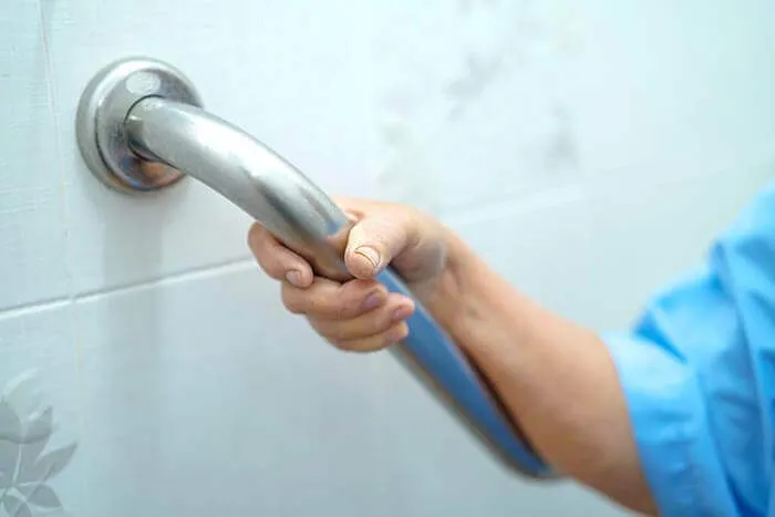 A person wearing a blue sleeve firmly grips a metal grab bar mounted on a tiled bathroom wall, providing support and stability.
