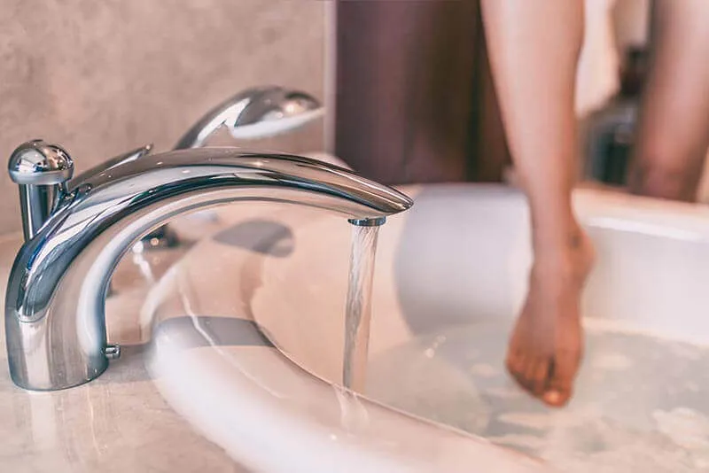 Chrome bath tap running water into a white bathtub with person's feet visible
