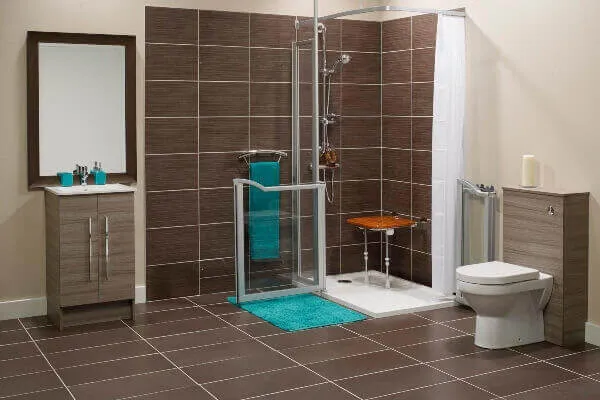 Modern accessible bathroom with a walk-in shower, wooden bench, glass doors, a toilet, a sink with a mirror, and teal bath mats and towels. Walls and floors have brown and beige tiles.
