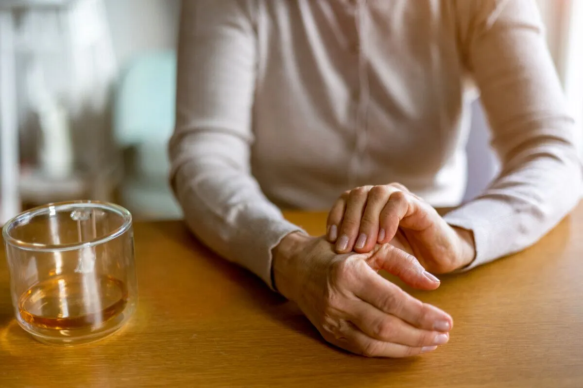 An older adult sits at a wooden table, holding their wrist with one hand as if in pain or discomfort. A glass of water is on the table nearby. The person is wearing a light-colored sweater.