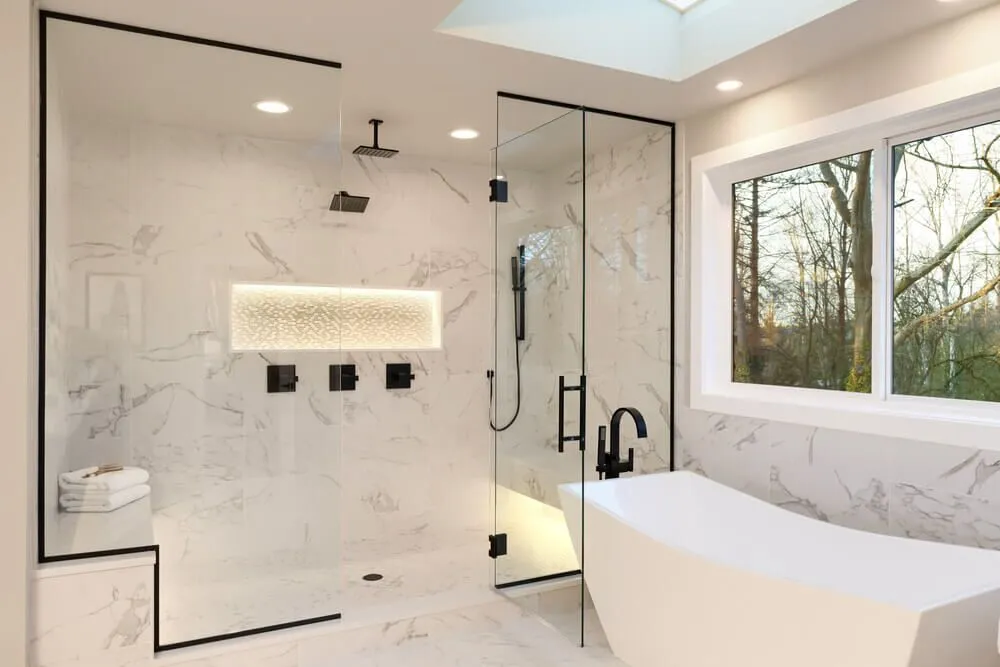 Modern bathroom with white marble walls and floor, a glass-enclosed walk-in shower with black fixtures, a built-in bench, a freestanding white bathtub, and a large window with a view of trees outside.