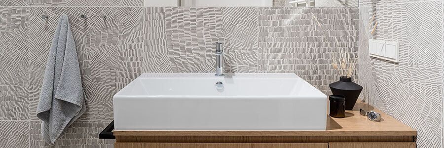 Modern bathroom sink with a square white basin on a wooden vanity, patterned light gray tile wall, a gray towel hanging on hooks, and a black vase with reed sticks and small glass container on the countertop.