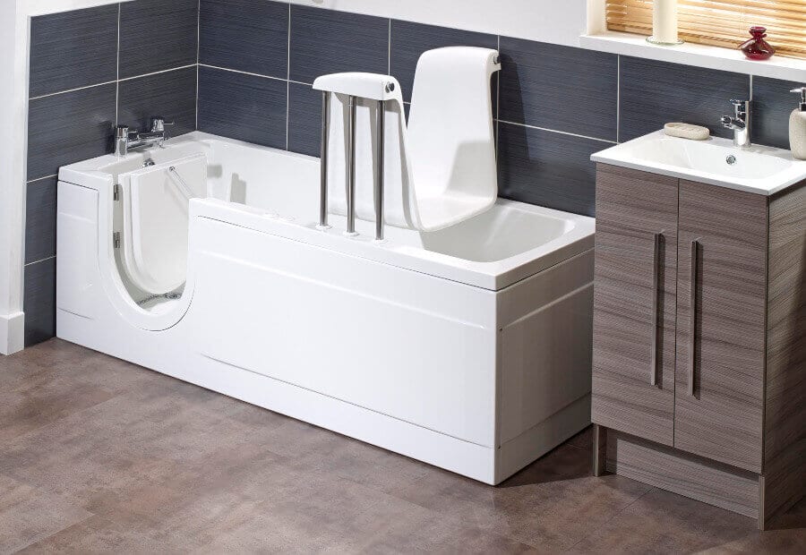 A modern bathroom with a walk-in bathtub featuring a door, a high-backed seat with armrests, and a grey vanity unit with a sink, set against dark tiled walls and a light brown floor.