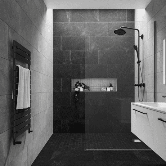 A modern bathroom with dark gray tiles, a walk-in shower featuring a black rainfall showerhead, a glass partition, a recessed shelf with toiletries and a plant, a heated towel rail, and a white vanity with a mirror.