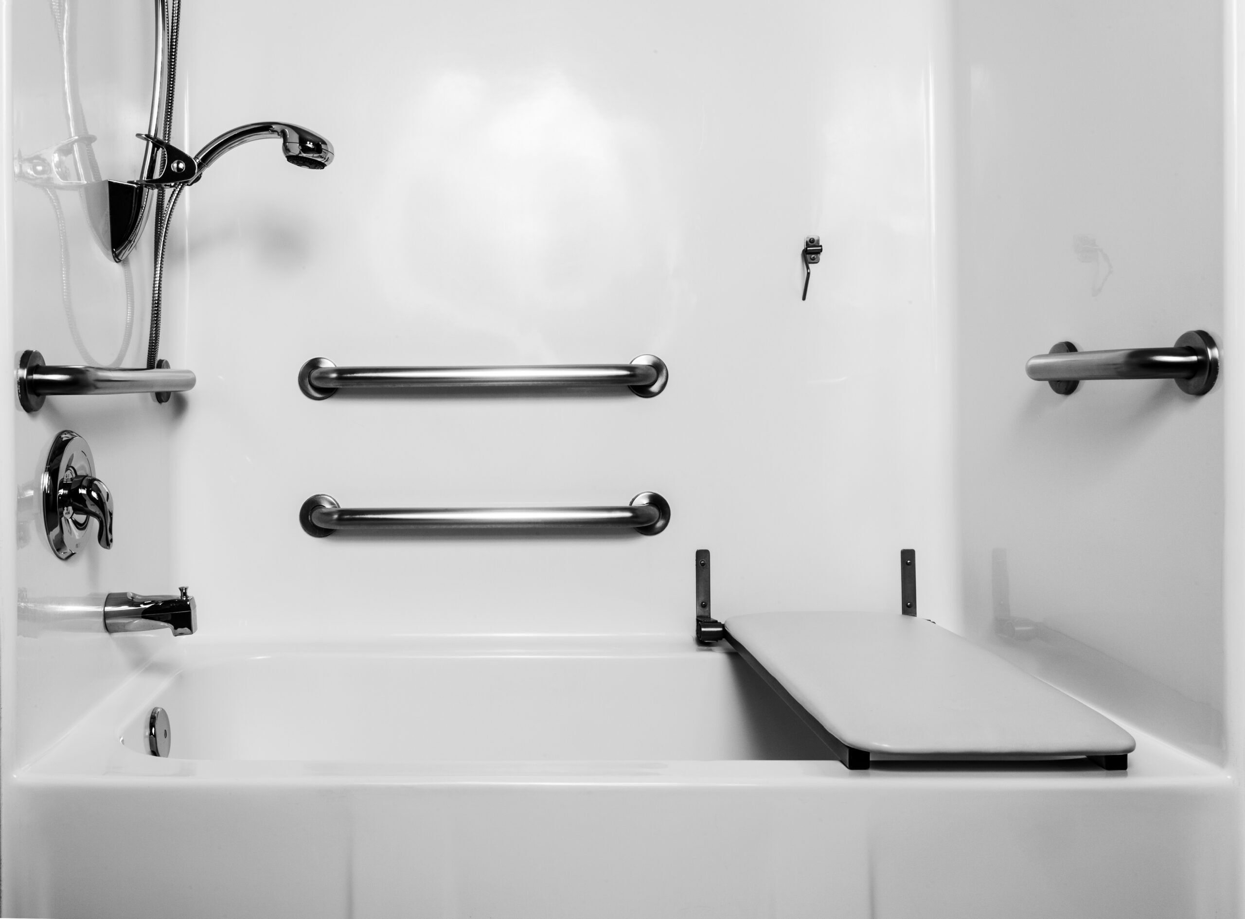 Accessible bathtub with a handheld showerhead, three horizontal grab bars, and a transfer bench on the right side, all within a white, tiled bathroom setting.