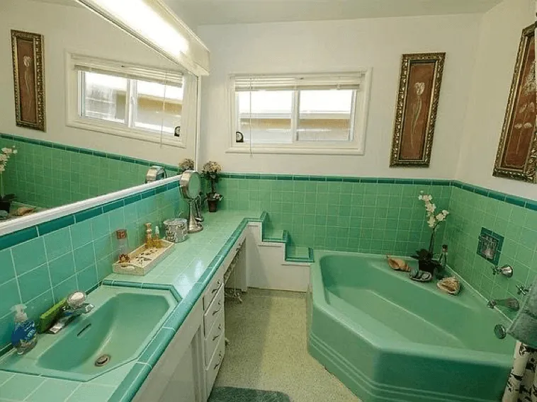 A bathroom with bright turquoise tiles, a matching hexagonal bathtub and sink, a large mirror, and two framed flower artworks on the white walls. The counter and bathtub have decorative items placed on them.