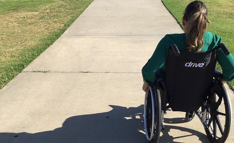 A woman with brown hair in a ponytail, wearing a green shirt, sits in a wheelchair on a paved path in a park, heading toward an open area with grass on each side.