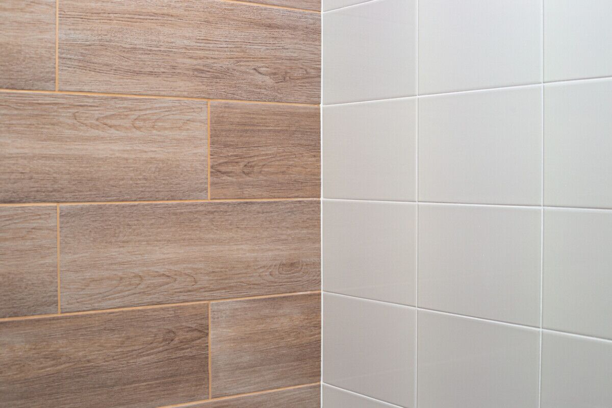 Close-up of two intersecting tiled walls; one wall has rectangular tiles with a woodgrain pattern, while the other wall has plain, light-colored rectangular tiles.