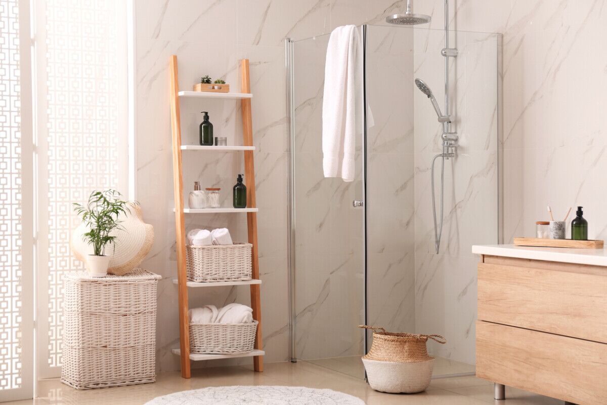 Modern bathroom with a glass shower, wooden ladder shelf holding towels and toiletries, wicker baskets, a potted plant, and light wood cabinetry against marble-patterned walls.