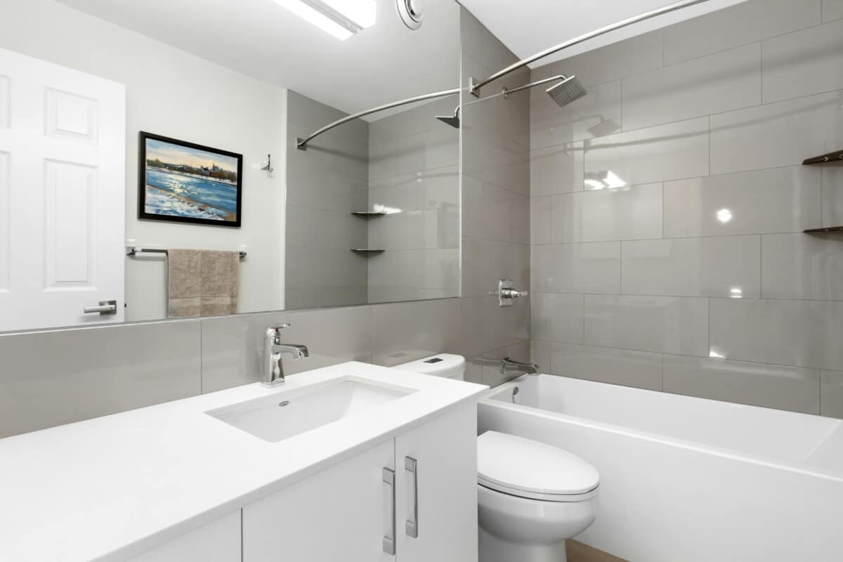 Modern bathroom with a white vanity, sink, and countertop, a large wall mirror, a toilet, and a bathtub with a curved shower rod. Light gray tiles line the shower wall; a painting hangs above a towel rack.