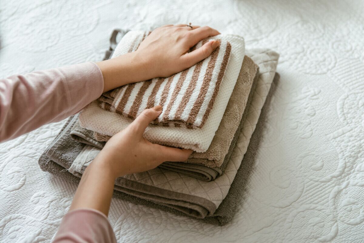 Hands neatly stacking a pile of folded towels and washcloths, including a brown and white striped towel, on a white textured bedspread.