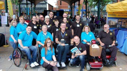 A group of people, some in wheelchairs, pose and smile together outdoors under a decorative gazebo. Some wear matching blue shirts, others wear black shirts, with event tents and banners visible in the background.
