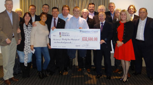 A group of people stand together indoors, smiling as they hold a large ceremonial check for ¬£32,600 made out to Help for Heroes from Premier Care.