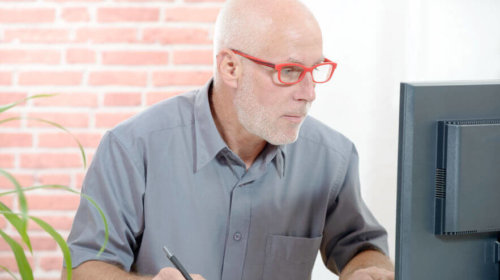 An older man with white hair and a beard, wearing red glasses and a gray shirt, sits at a desk, looking intently at a computer monitor while holding a pen and taking notes.