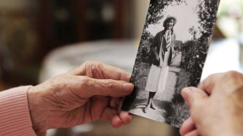 An elderly person with wrinkled hands holds a black-and-white photo of a young woman wearing a hat and coat, standing outdoors on a path lined with trees.