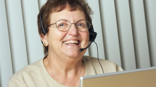An older woman wearing glasses and a headset smiles while sitting in front of a laptop, with vertical blinds in the background.