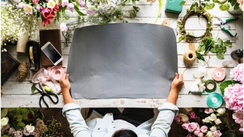A person sits at a wooden table covered with flowers, ribbons, and craft supplies, holding a large blank sheet of black paper in the center, ready for a floral or creative project.