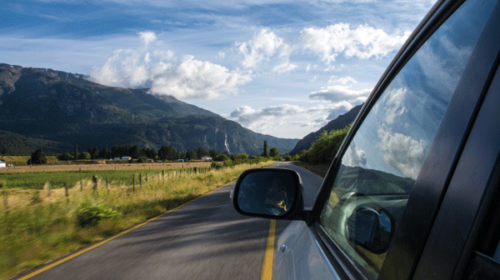 A car drives along a rural road with mountains, green fields, and clouds visible in the distance. The cars side mirror and window reflect the scenic landscape.