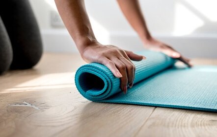 Close-up of a person‚Äôs hands rolling up a blue yoga mat on a wooden floor, with natural light coming from the background.