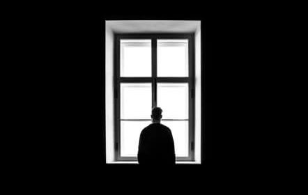 A person stands in silhouette, facing a large window with four panes, surrounded by darkness, looking outside.