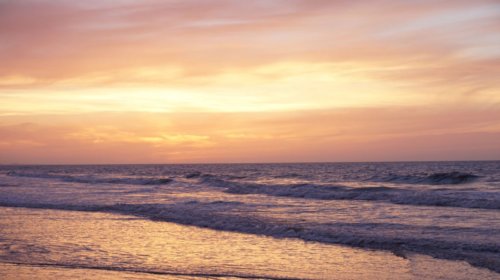 Waves gently roll onto a sandy beach under a vibrant sunset sky, with shades of pink, orange, and yellow reflecting on the water and horizon.