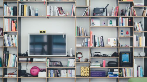 A modern bookshelf filled with books, small figurines, electronics, and various decorative items surrounds a flat-screen TV at the center. The shelves are neatly organized and the floor is wooden.
