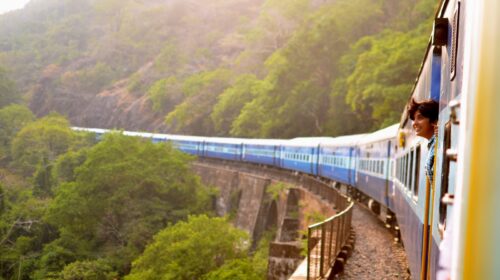 A blue train winds through lush green hills, crossing a bridge. A smiling person leans out of a window, enjoying the scenic view. The landscape is sunlit and vibrant.