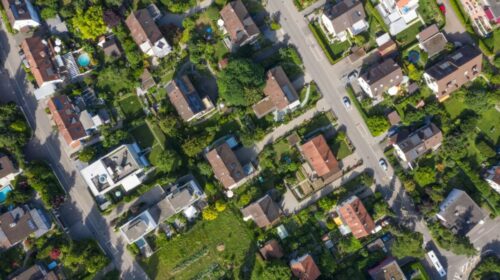 Aerial view of a suburban neighborhood with houses, green lawns, gardens, and tree-lined streets intersecting the area.