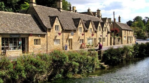 Row of charming stone cottages with sloped roofs along a riverbank, lush greenery in the foreground, and a person sitting by the water in a scenic village setting under a partly cloudy sky.