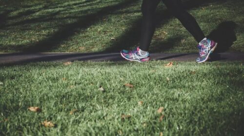 A person wearing black leggings and colorful running shoes walks on a paved path through a grassy area, with sunlight casting long shadows on the grass.