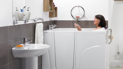 A woman relaxes in a modern walk-in bathtub in a bathroom with gray and white tiles, a wall-mounted sink, and shelves holding toiletries and towels.