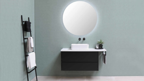 A modern minimalist bathroom with pale green walls, a large round mirror, a black vanity with a white sink, a small plant, and a black towel ladder holding folded towels.