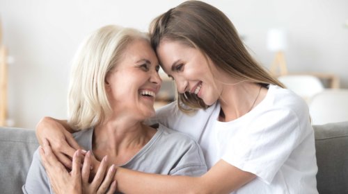 Two women, one older with gray hair and one younger with long brown hair, sit close together on a couch, smiling and embracing each other affectionately in a bright, cozy room.