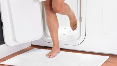 Person stepping barefoot out of a white walk-in bath door onto a white bath mat