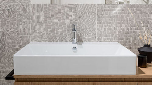 Modern bathroom sink with a square white basin on a wooden vanity, patterned light gray tile wall, a gray towel hanging on hooks, and a black vase with reed sticks and small glass container on the countertop.