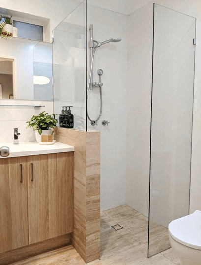 A walk in shower next to a wooden storage unit sink, with a plant 