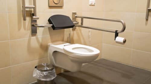 A clean, accessible restroom with a wall-mounted toilet, grab bars, a black backrest, a wall-mounted toilet paper holder, and a small trash can with a liner on a tiled floor and beige tiled walls.