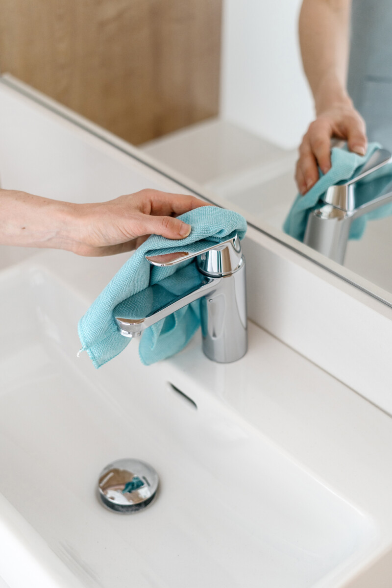 Vertical shot of a cleaning and polishing metal water tap with a blue microfibra rag in the bathroom. White ceramic washbasin and big mirror near the faucet