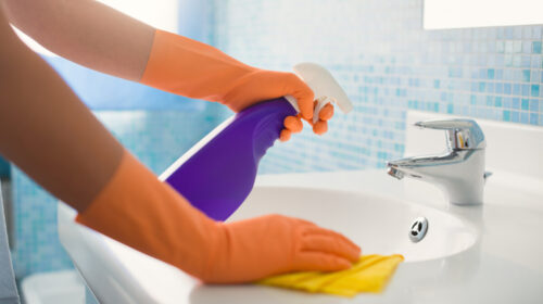 woman doing chores in bathroom at home, cleaning sink and faucet with spray detergent. Cropped view